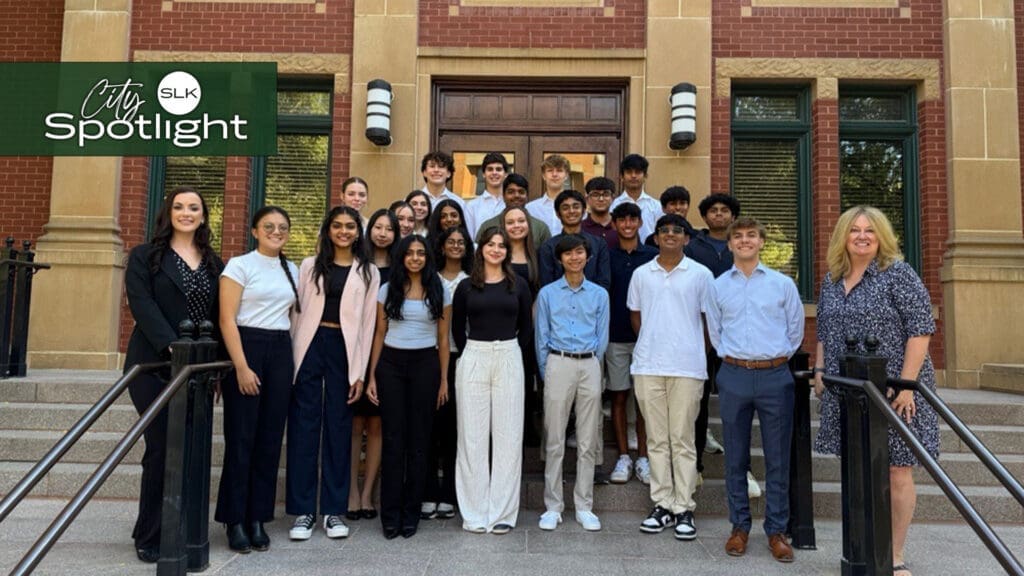 A group of students and a city staff member, and a city official stand on steps in front of a brick building, smiling. The group is dressed in business casual attire, with some wearing formal outfits. The building features tan stone trim and decorative star details.